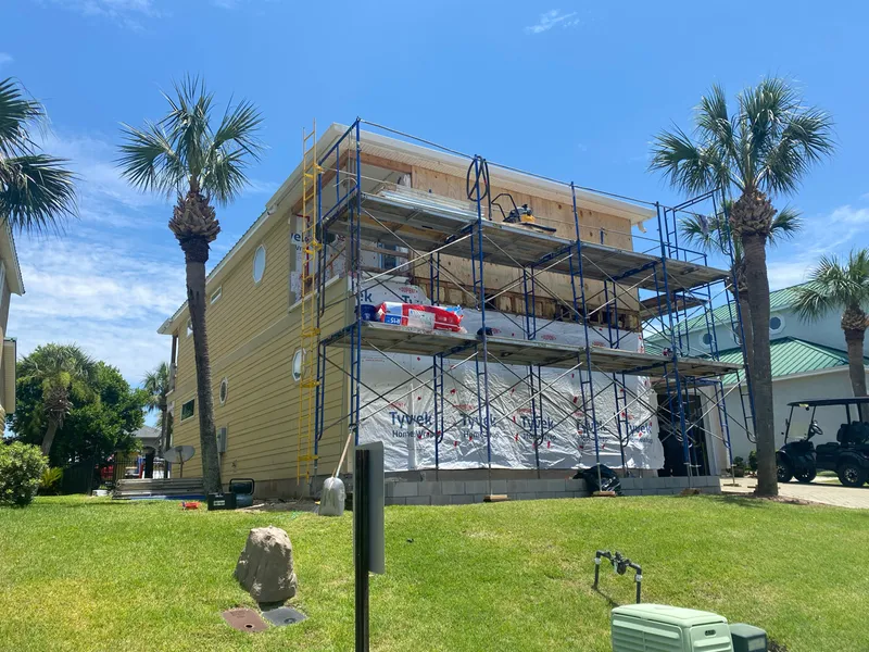 The house stripped to bare sheathing and housewrap, surrounded by scaffolding, with new siding started on the left elevation.