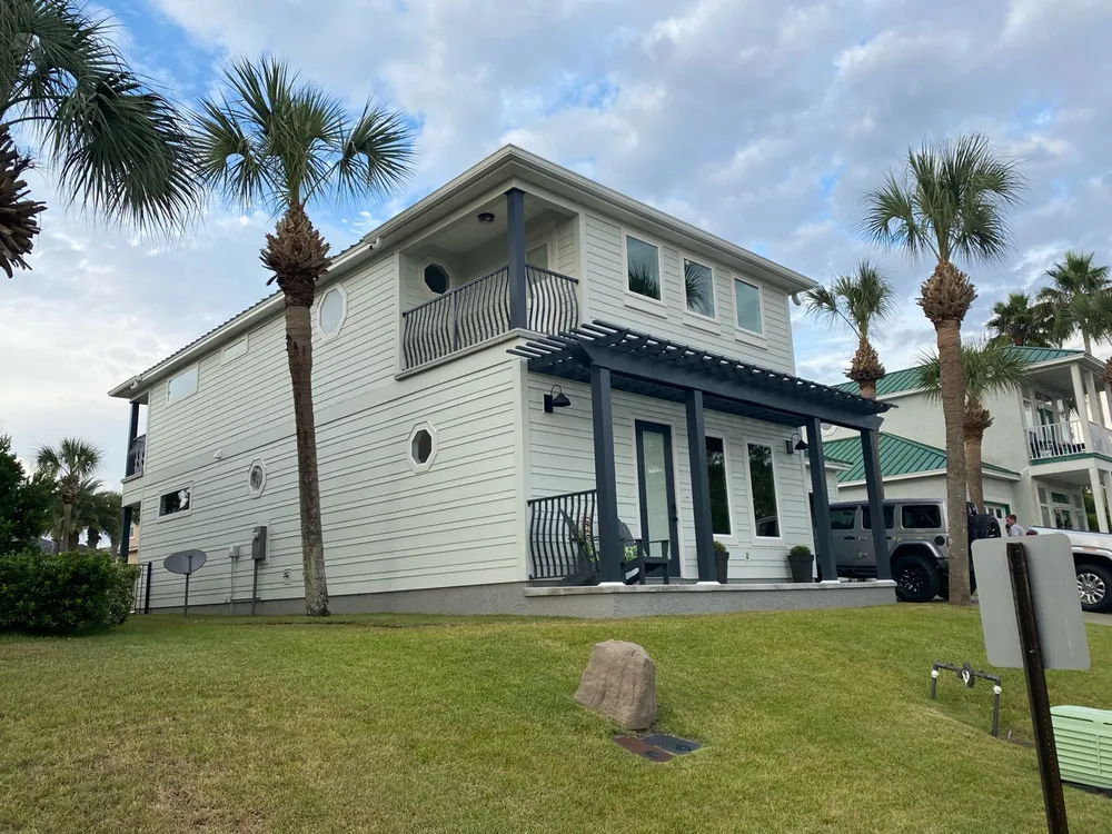 A completed two-story home with pale siding, a dark pergola over the front porch, and palms in the foreground.