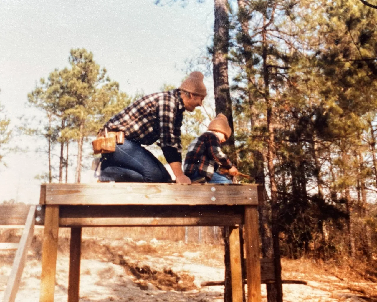 A 1982 photograph of Guy Garrett's father kneeling on a framed floor with Guy as a small boy beside him holding a hammer, among pine trees.