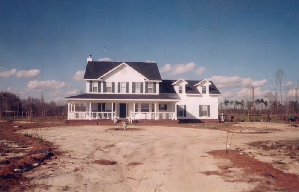 A two-story white farmhouse with a wraparound porch and dormers, standing on open farmland.