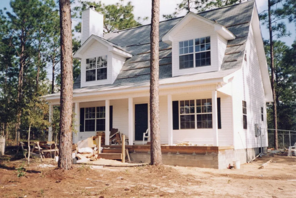 A white story-and-a-half house with dormers and a deep front porch, standing among pines.