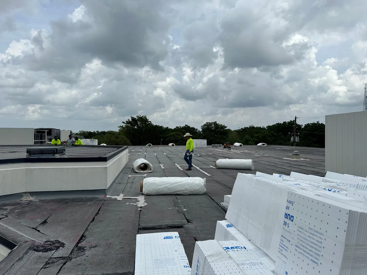 Insulation boards stacked on the roof in the foreground, with a crew member and membrane rolls beyond.