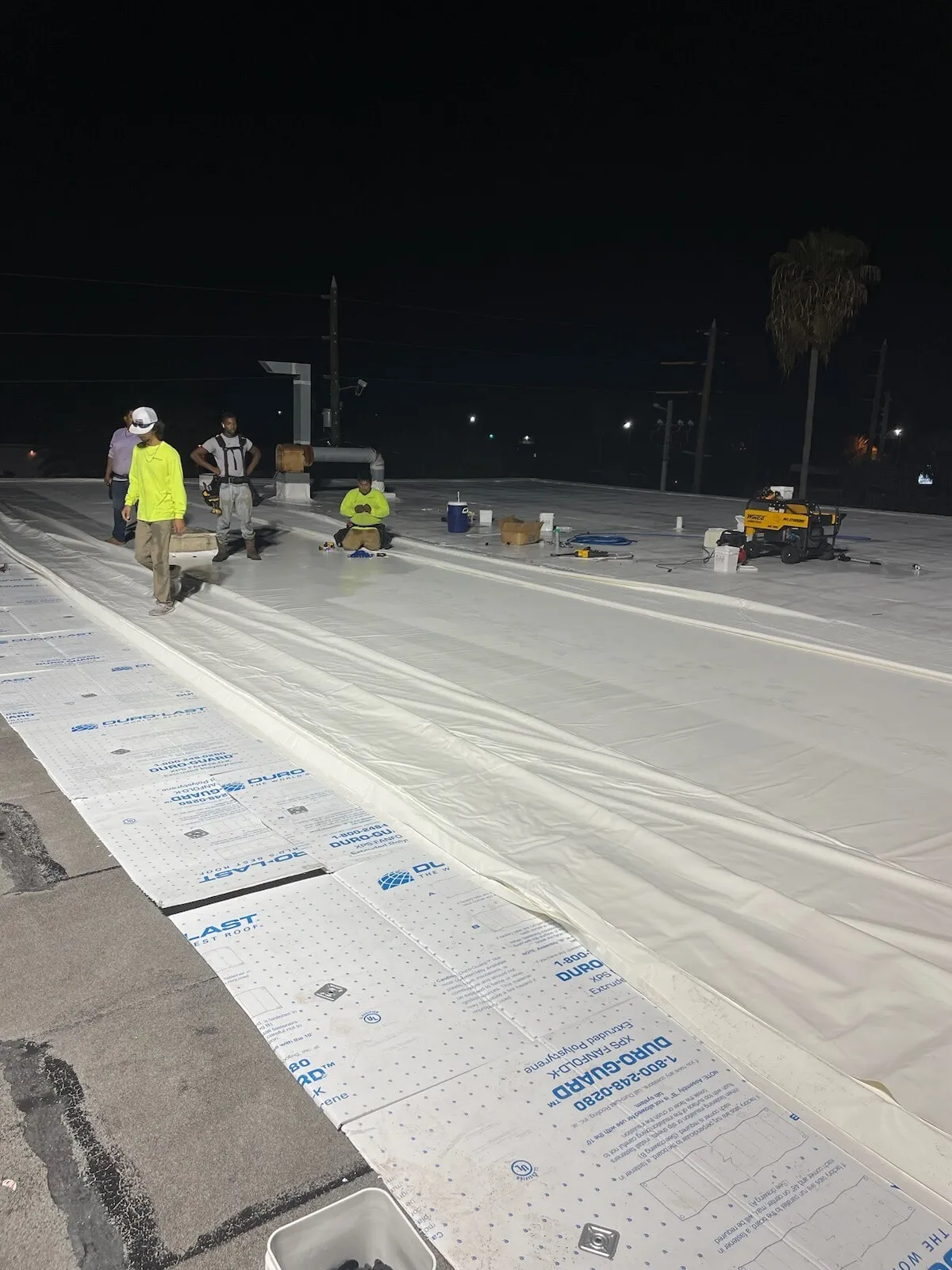 Crew members in safety vests working at night on white membrane laid over printed insulation boards, under work lights.