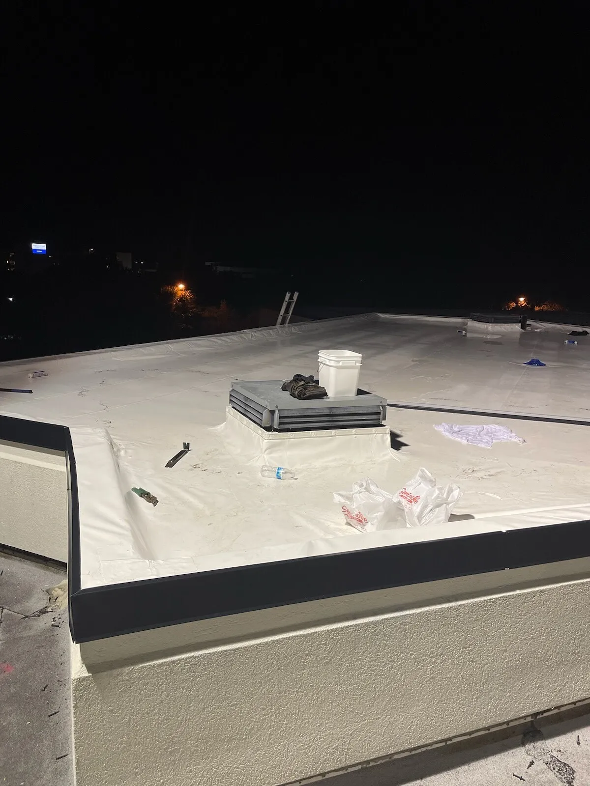 A raised curb and the roof edge at night, with membrane, tools, and materials in the work area.