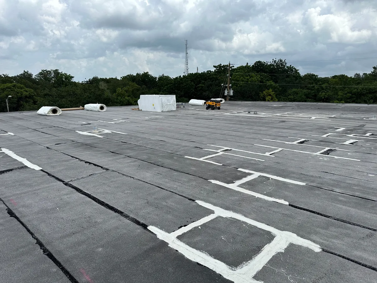 Broad existing roof surface with white markings, membrane rolls, and staged materials in the distance.