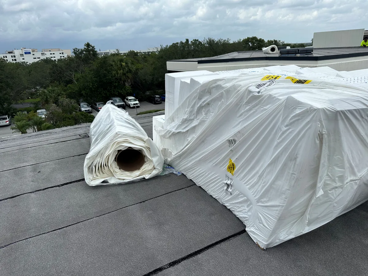 Wrapped stacks of insulation boards and a membrane roll staged on the existing roof near the roof edge.