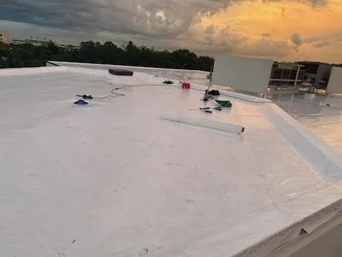 White membrane across a broad roof area at dusk, with tools and a cord still on the roof and rooftop equipment beyond.