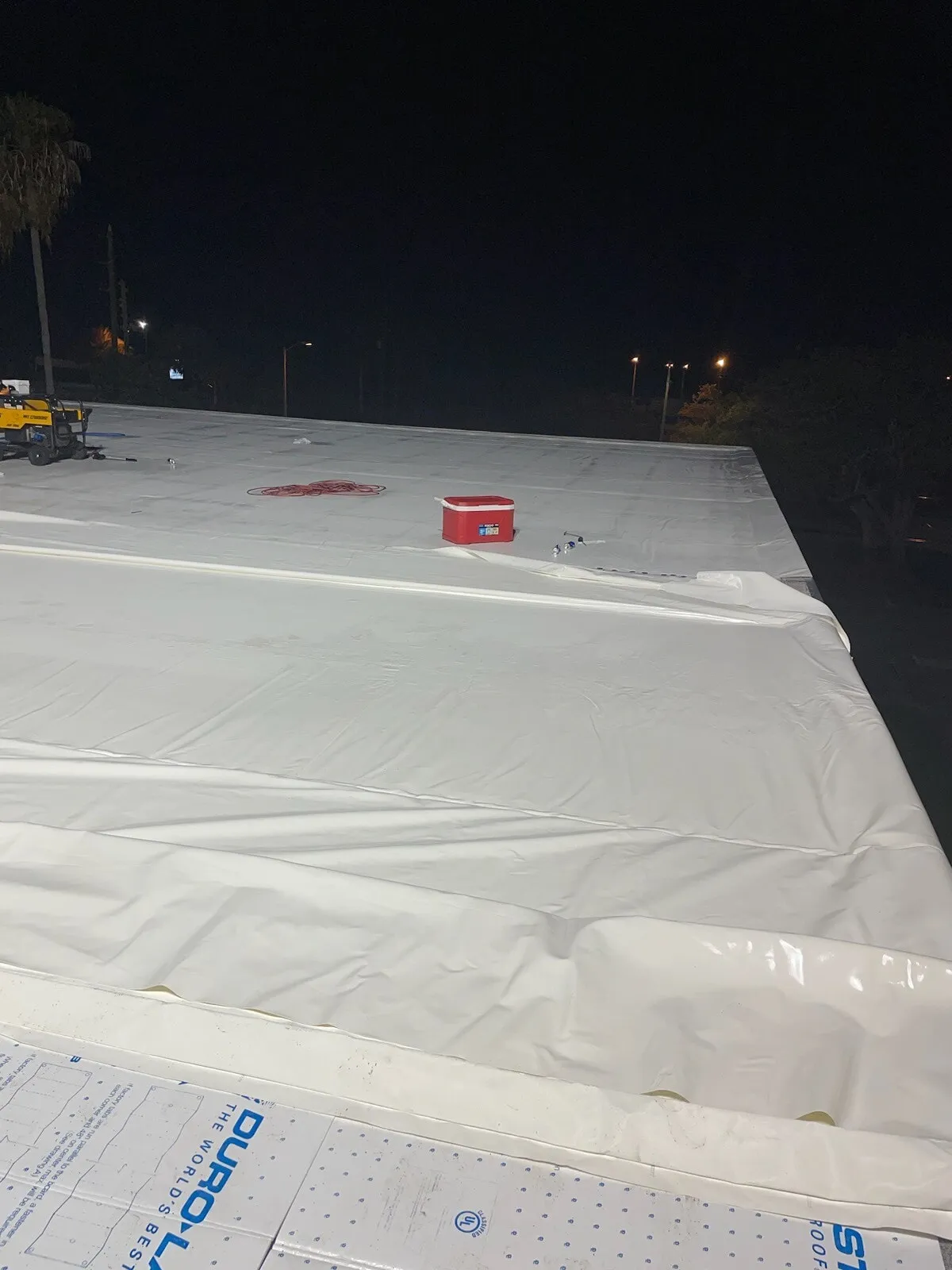 White membrane laid across the roof over insulation boards at night, with loose edges still to be completed.