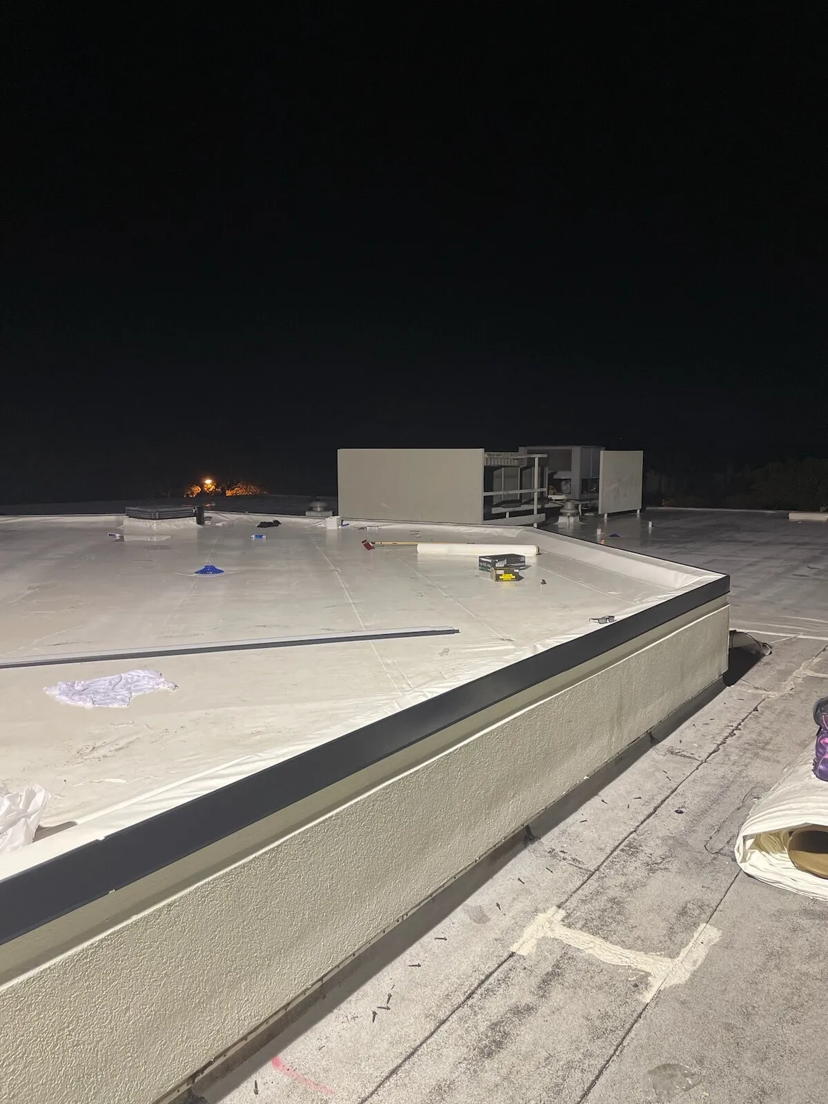 Roof perimeter at night where new white membrane meets a lower roof level along a parapet edge.