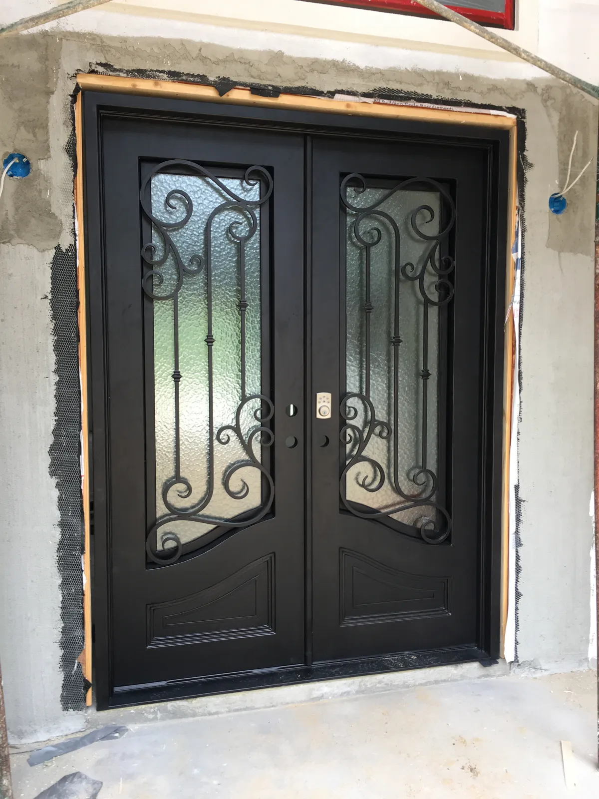 Dark double entry doors with textured glass and scrollwork, hung in the opening, with unfinished stucco and protective tape around them.
