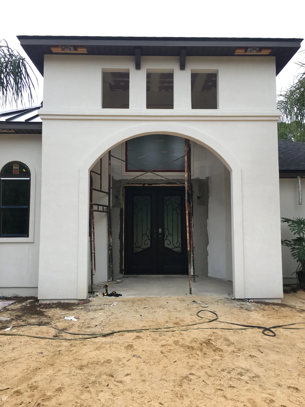 Stucco front entry with a tall arched opening, three small upper windows, and dark double doors, with scaffolding and bare sand in front.