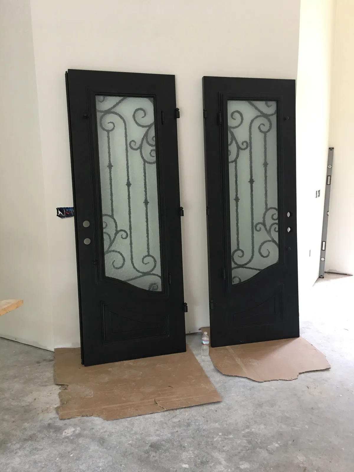 Two dark steel door slabs with scrolled glass panels leaning against an interior wall on cardboard, before hanging.
