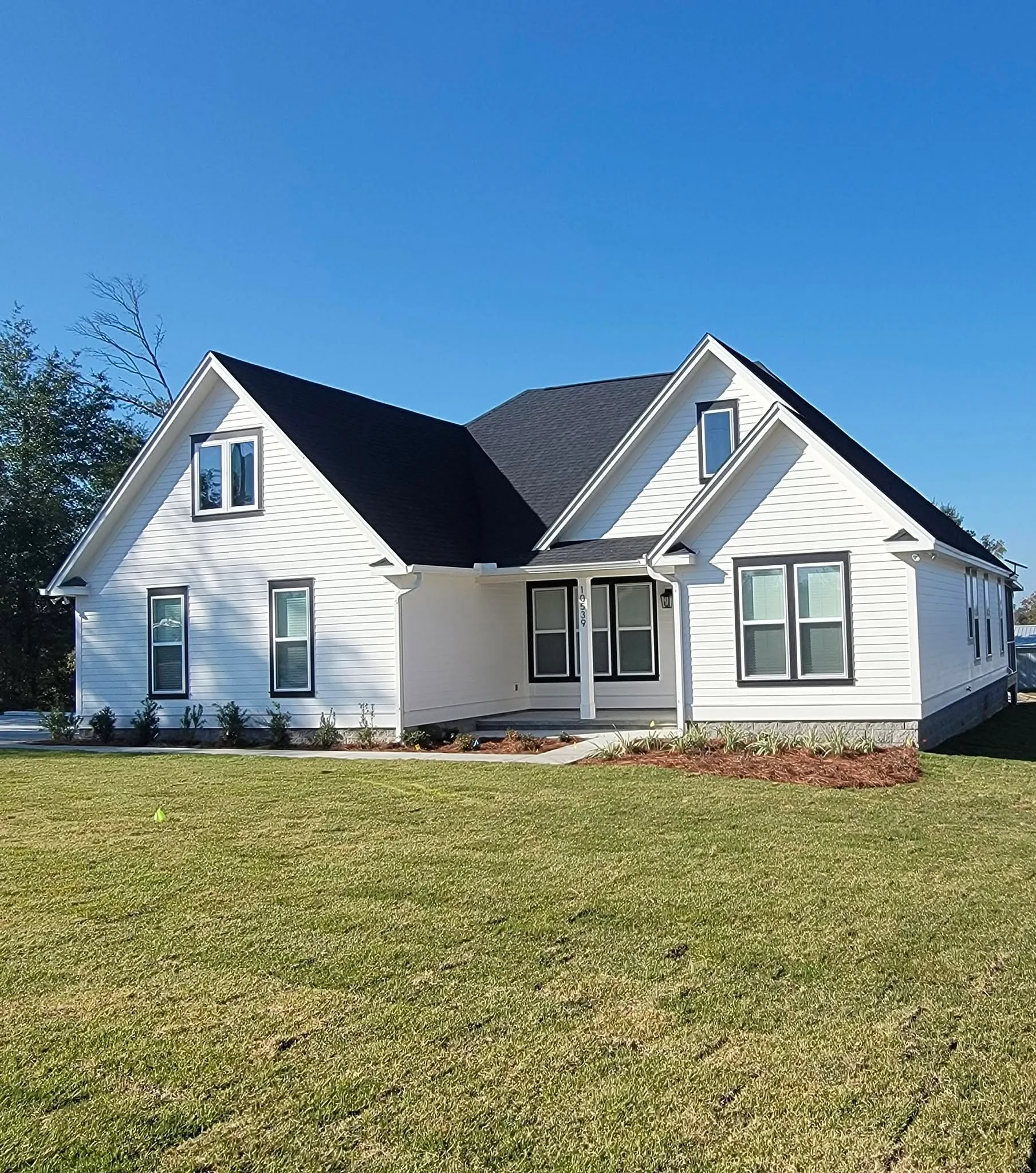 Completed white custom home with a dark roof and trim, steep front gables, planting beds, and a green lawn.
