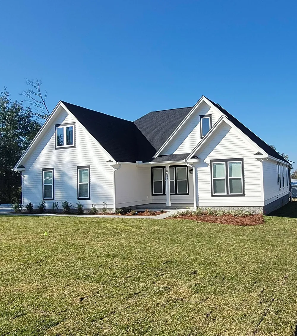 Completed white custom home with a dark roof and trim, steep front gables, planting beds, and a green lawn.
