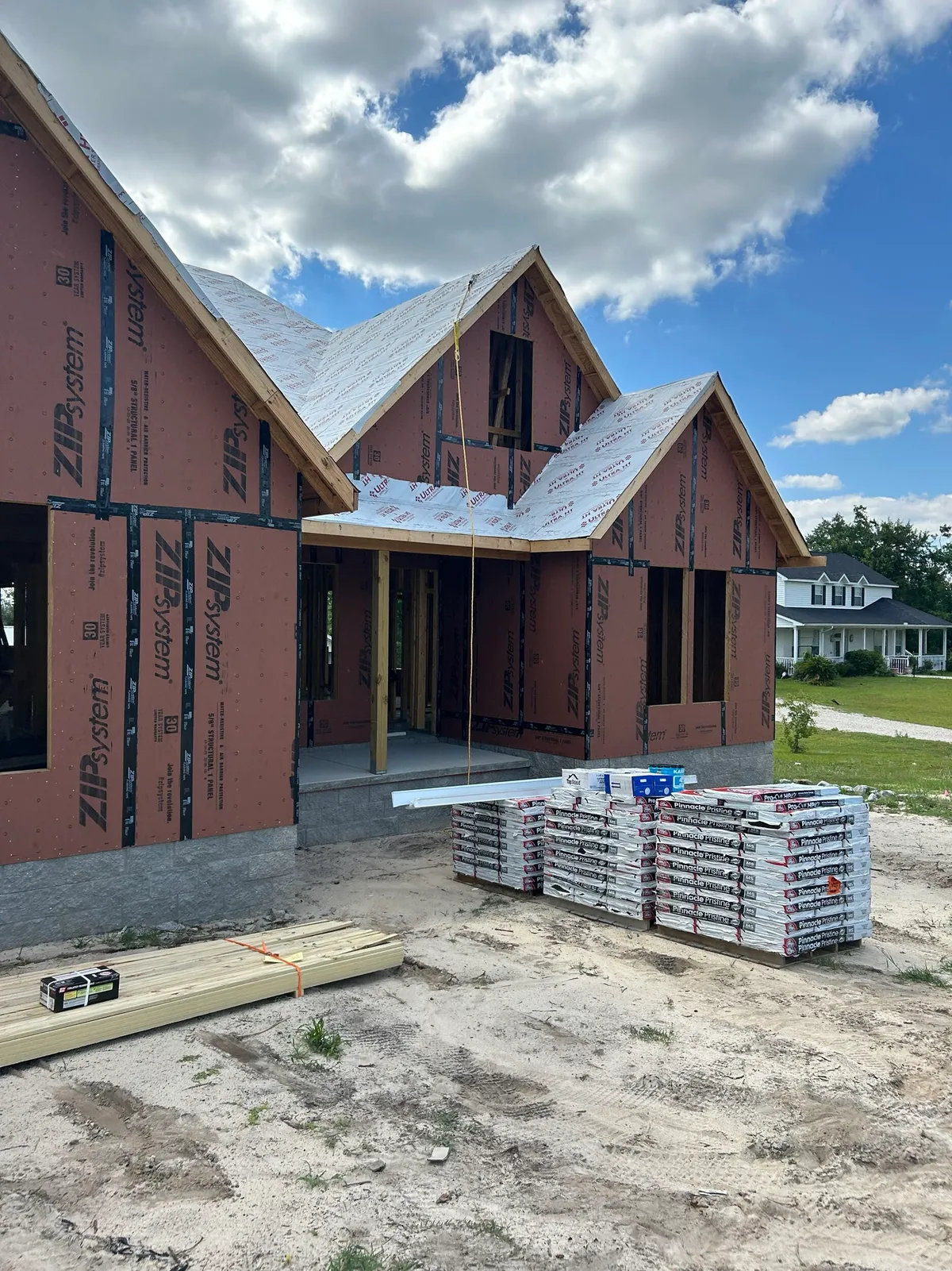 Front of the sheathed house with roof underlayment on and bundles of shingles stacked in front.