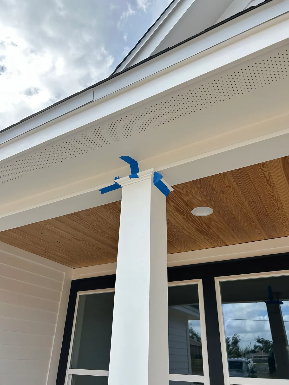 White porch column with painter’s tape at its top, below a vented soffit and wood ceiling.