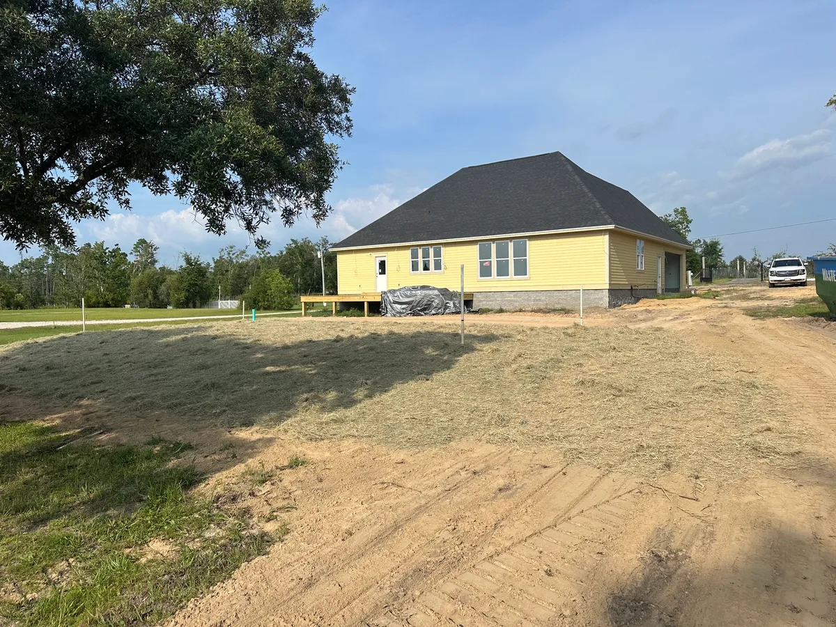 Distant rear view of the house with a shingled roof and unpainted yellow siding across a graded yard.