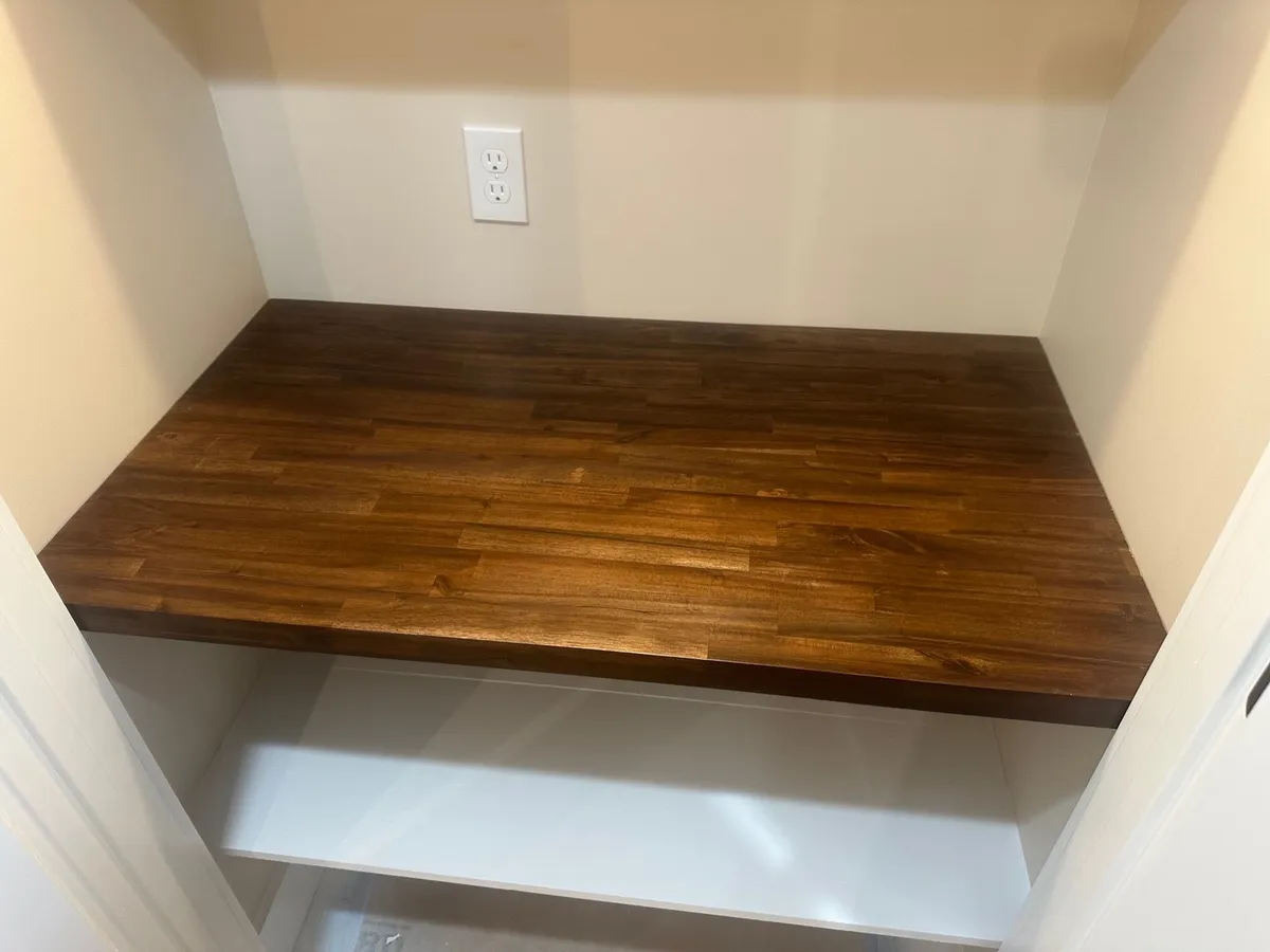 Dark wood countertop in a white built-in nook, with an outlet above and a shelf below.