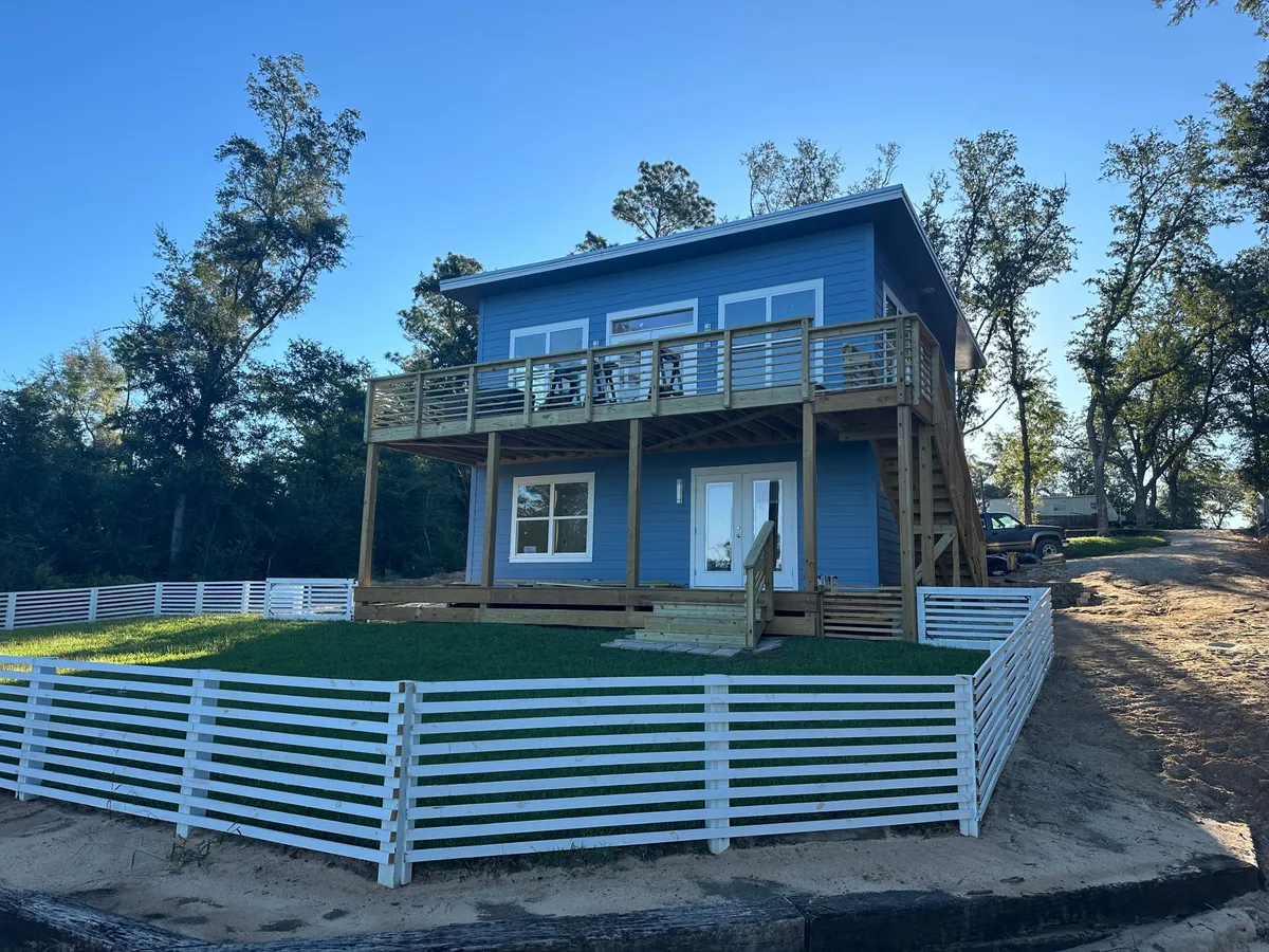 Lake-facing side of a blue two-story home with an upper deck, lower porch, and fenced lawn.
