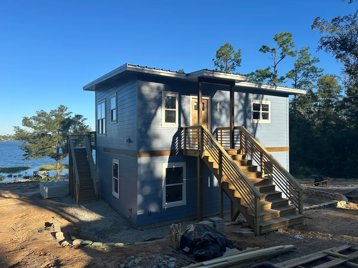 Two-story blue-sided home with wood exterior stairs to the upper entry, beside a lake, with site work still underway.