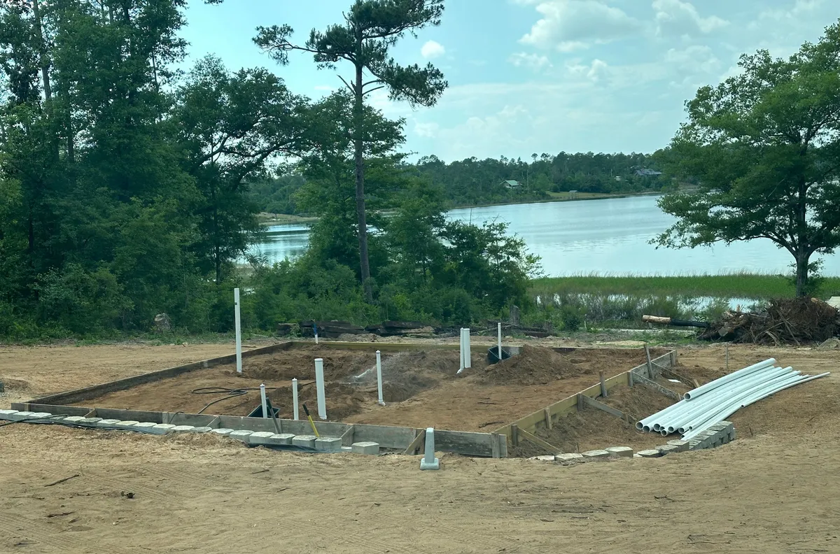 Foundation area with block edges and plumbing pipes rising from the ground, with the lake and trees beyond.