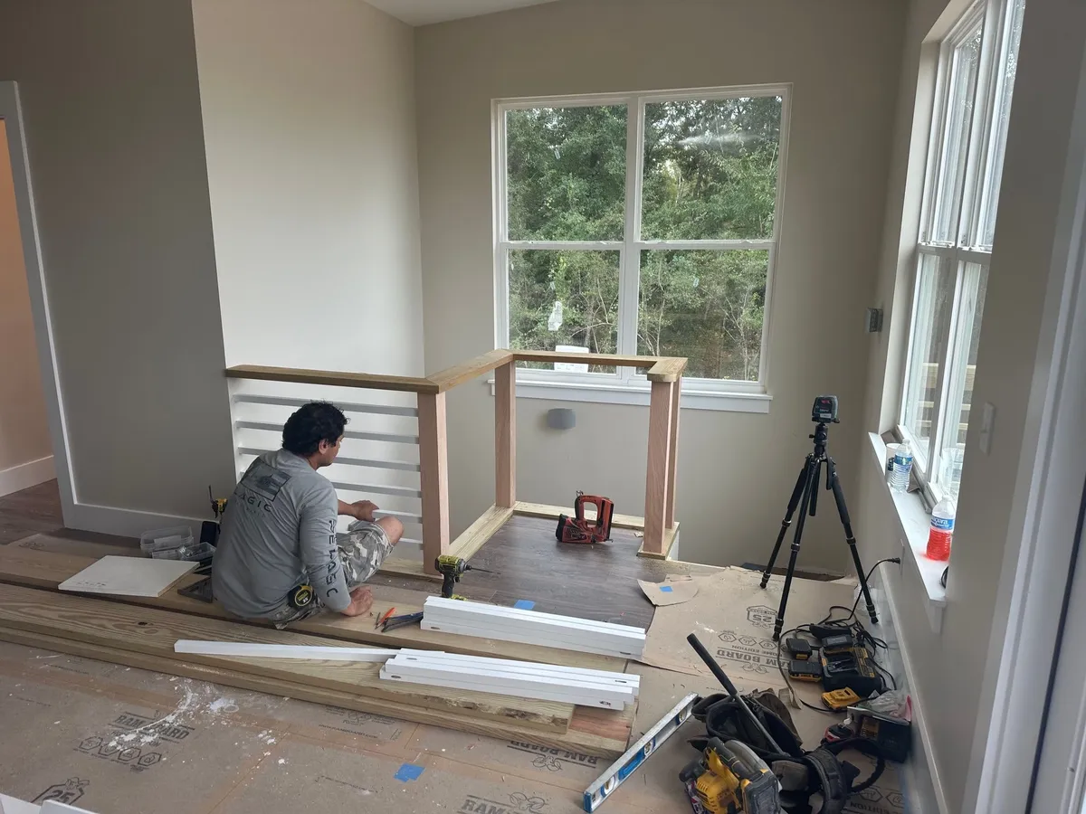 A worker assembling wood posts and white horizontal rails around the stair opening, with tools on the floor.