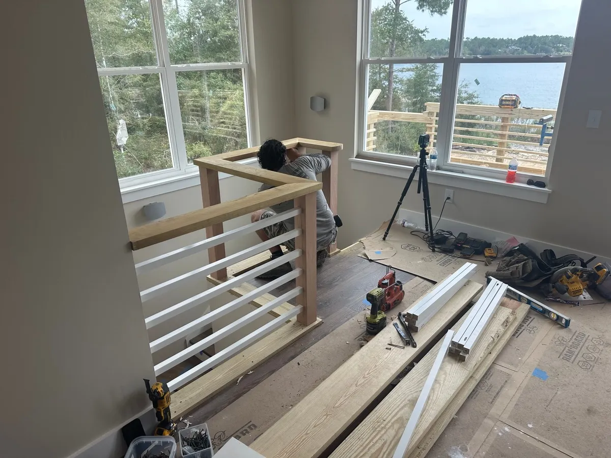 A worker fitting the wood railing over white horizontal rails at the stair opening, with a tripod-mounted level nearby.