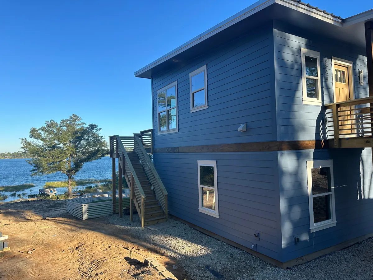 Side of the two-story blue home with an exterior stair and upper landing, and the lake beyond.