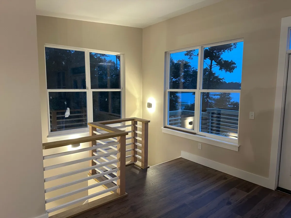Stair opening framed by wood posts and a wood cap with white horizontal rails, in a corner with two windows.
