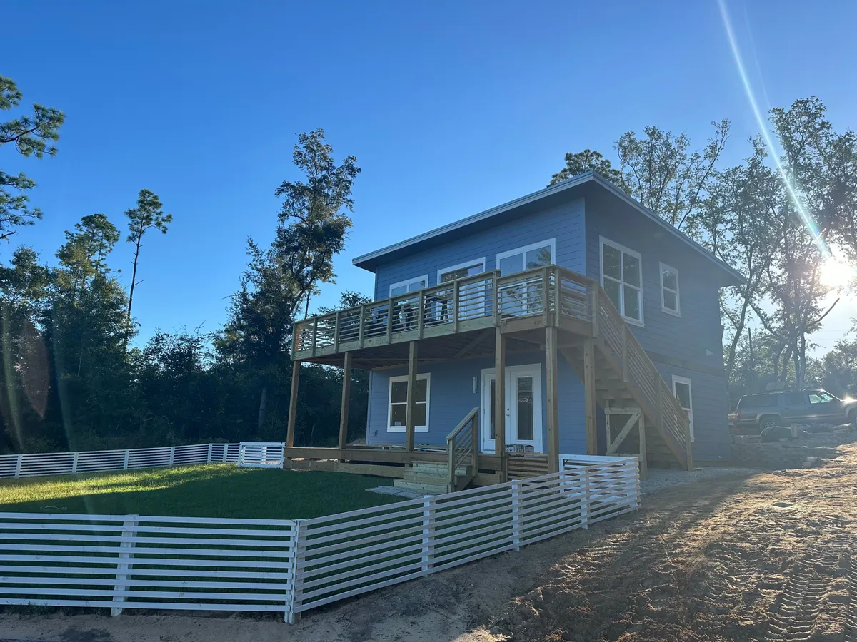 Lake-facing side of the home with an upper deck over a sheltered lower area, a new lawn, and white horizontal-board fencing.