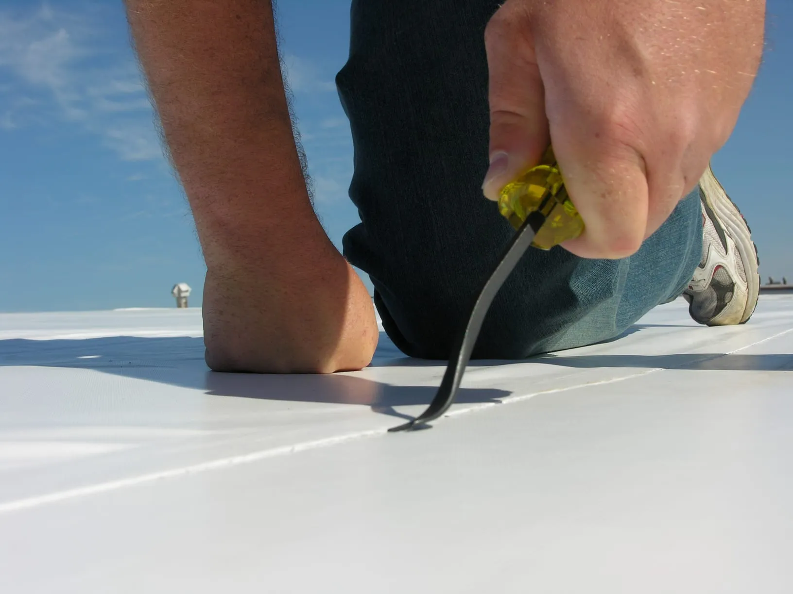 A person kneeling on a white membrane roof, probing a welded seam with a hand tool.