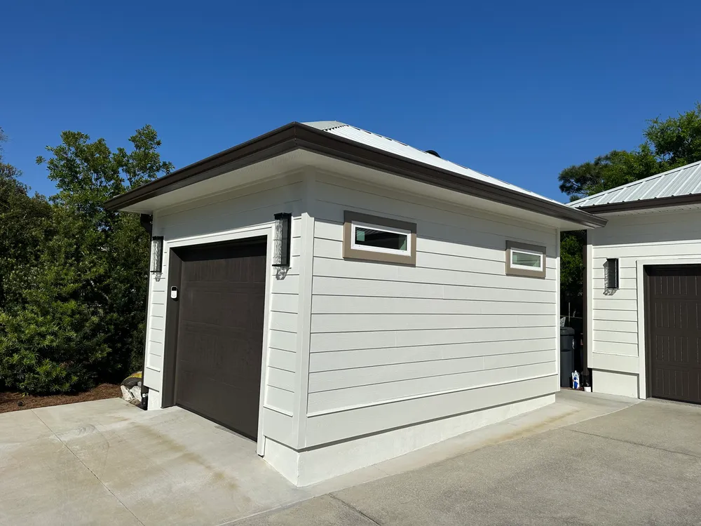 Completed detached golf cart garage with white lap siding, a metal hip roof, high windows, and a dark garage door beside the matching existing building.