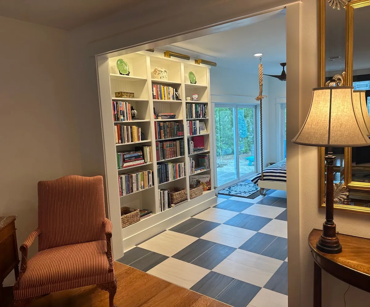 Bookcase and checkerboard tile floor seen from the living room, beside an armchair and a table lamp.