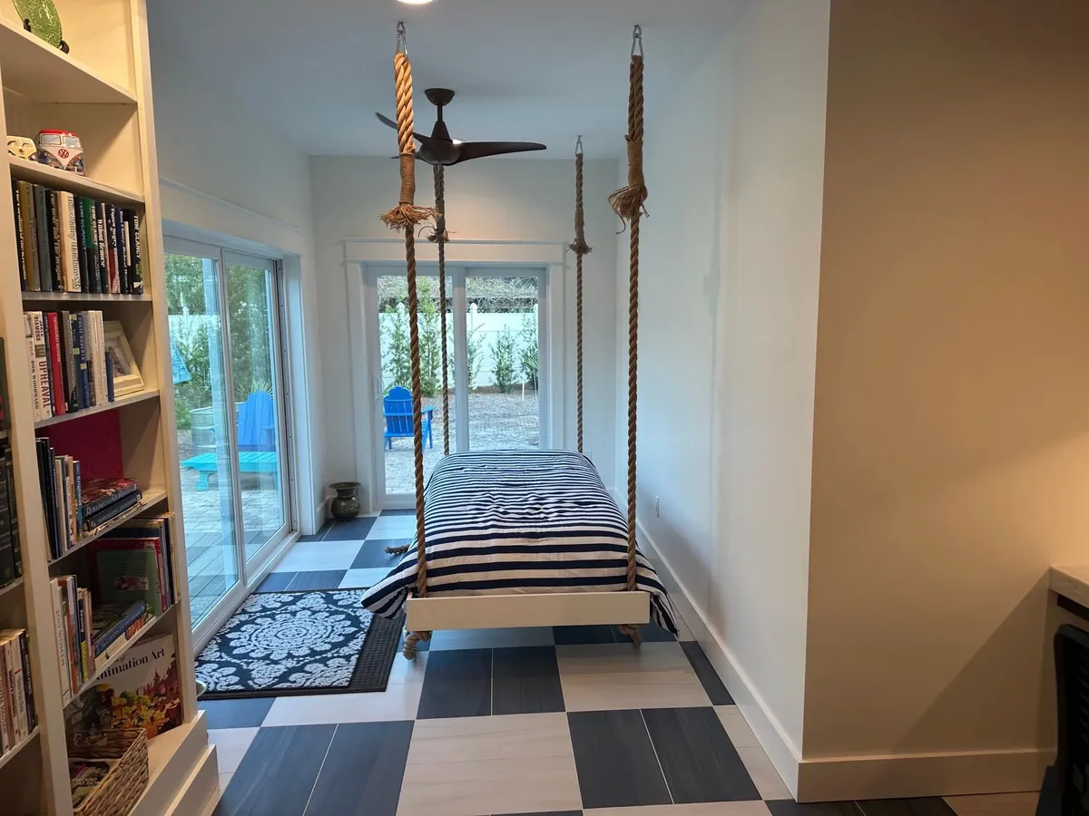 Hanging bed suspended on ropes beside sliding glass doors, with the bookcase at left and a ceiling fan above.