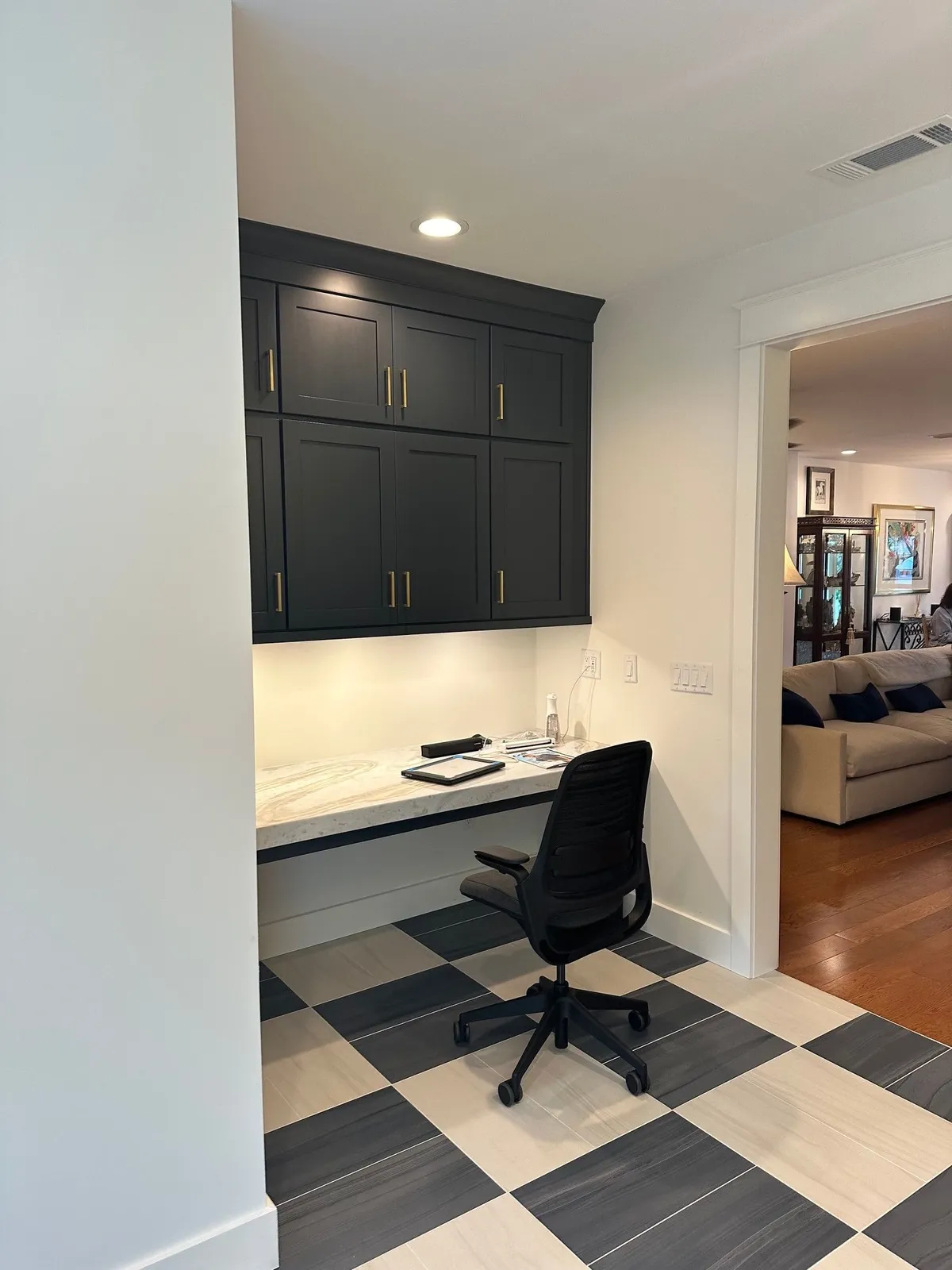 Office nook with dark upper cabinets over a white desk surface and a desk chair, beside an opening to the living room.