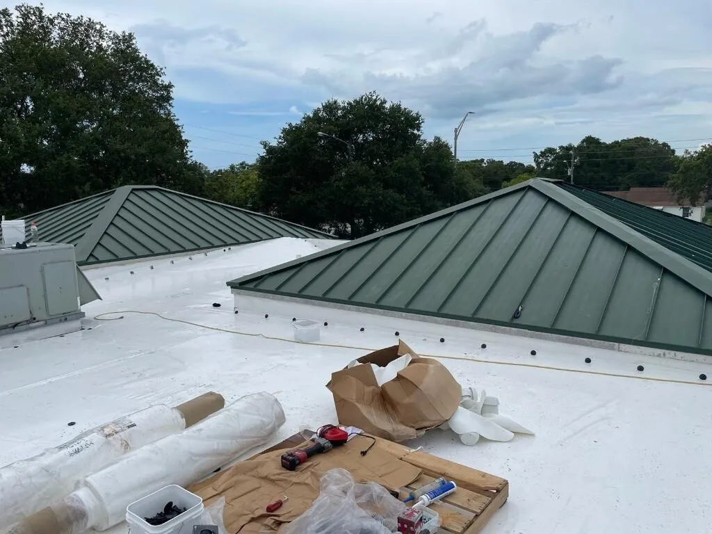 New white membrane beside green metal roof sections, with membrane rolls, tools, and packaging on the roof.