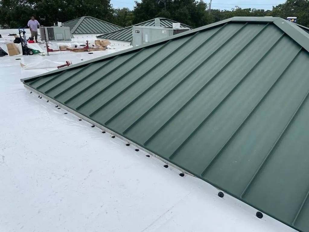 White membrane along the base of green standing-seam metal roof sections, with crew members and materials in the distance.