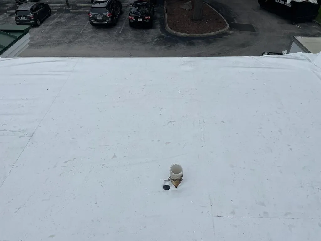 A pipe penetration through the white membrane near the roof edge, with a parking lot below.