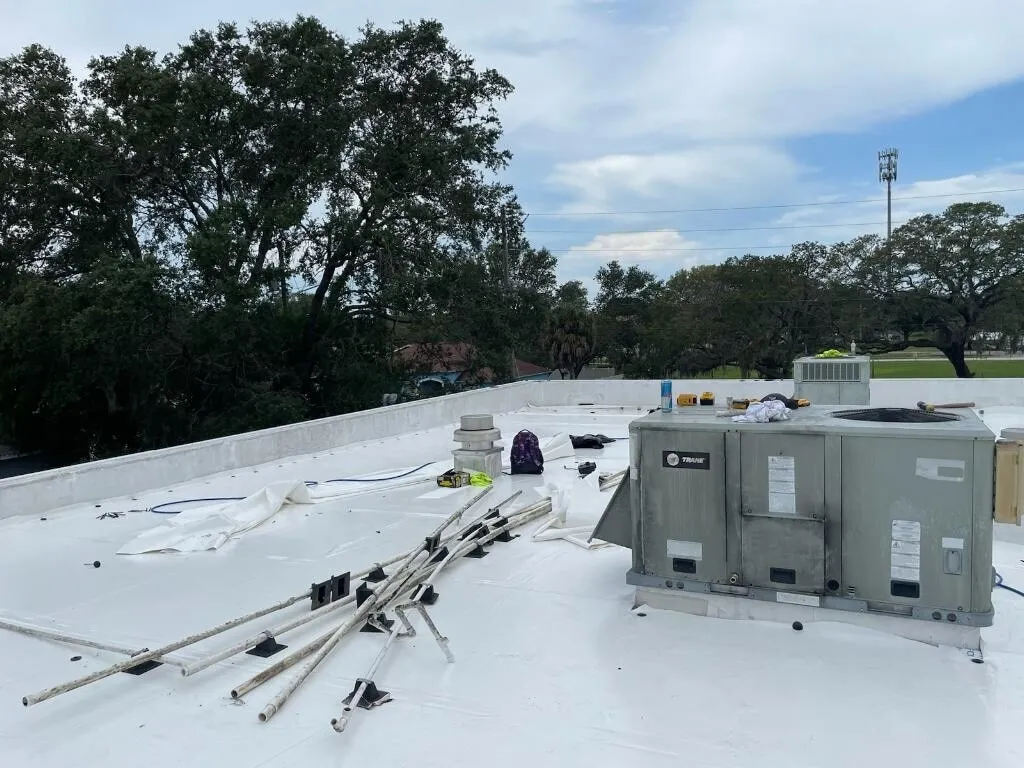 A rooftop unit on the white membrane, with pipes, tools, and materials still on the roof and trees beyond.