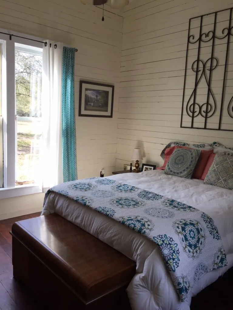 The furnished bedroom, with a made bed, wood walls, and daylight through the window.