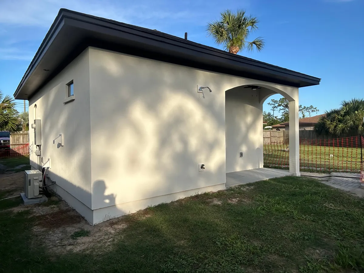 Side and rear of the white stucco studio, with the arched back porch, an air conditioning condenser, and orange safety fencing beyond the lawn.