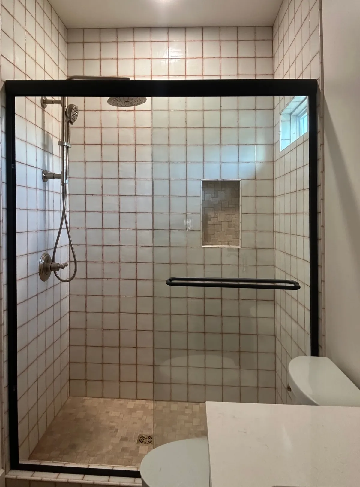 Shower with square white wall tile, a recessed niche, a small window, and a black-framed glass door, beside the toilet and vanity.