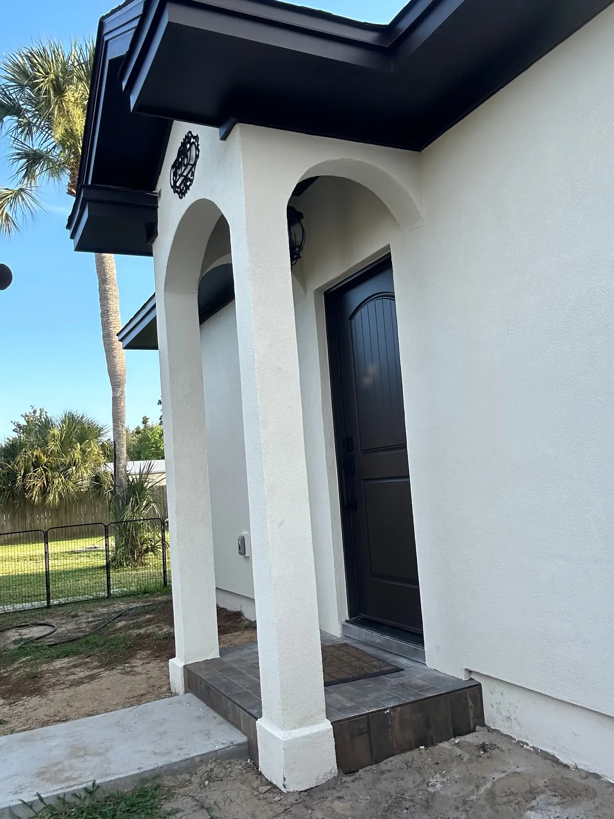 Angled view of the arched entry, with stucco columns, a dark door, a stone-look step, and dark fascia at the roofline.