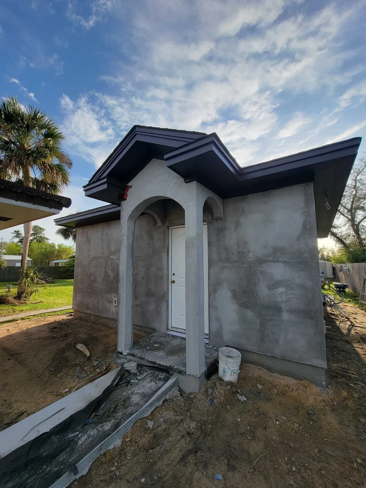 Front of the studio after stucco, with the arched entry, gable returns, and a white door, while site work continues.