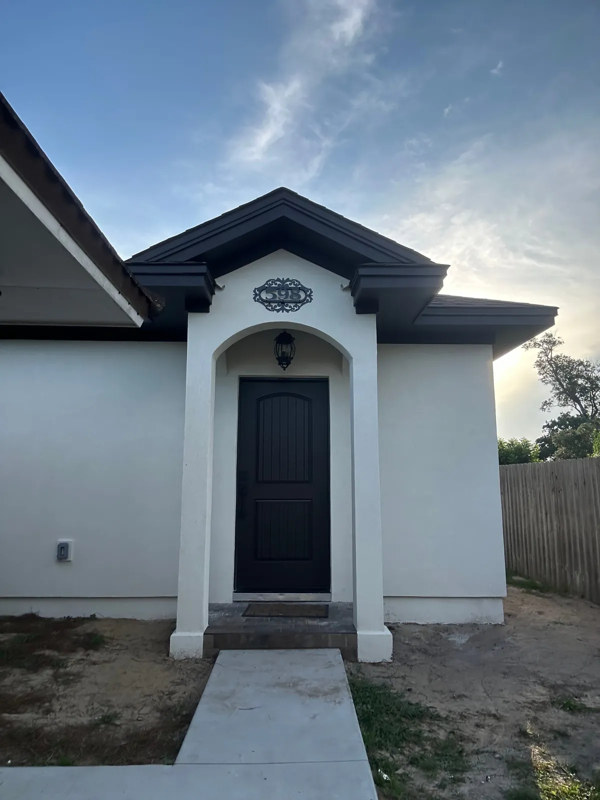 White stucco studio with an arched covered entry, a dark wood door, a hanging lantern, and gable returns at the entry roofline, with a concrete walk in front.