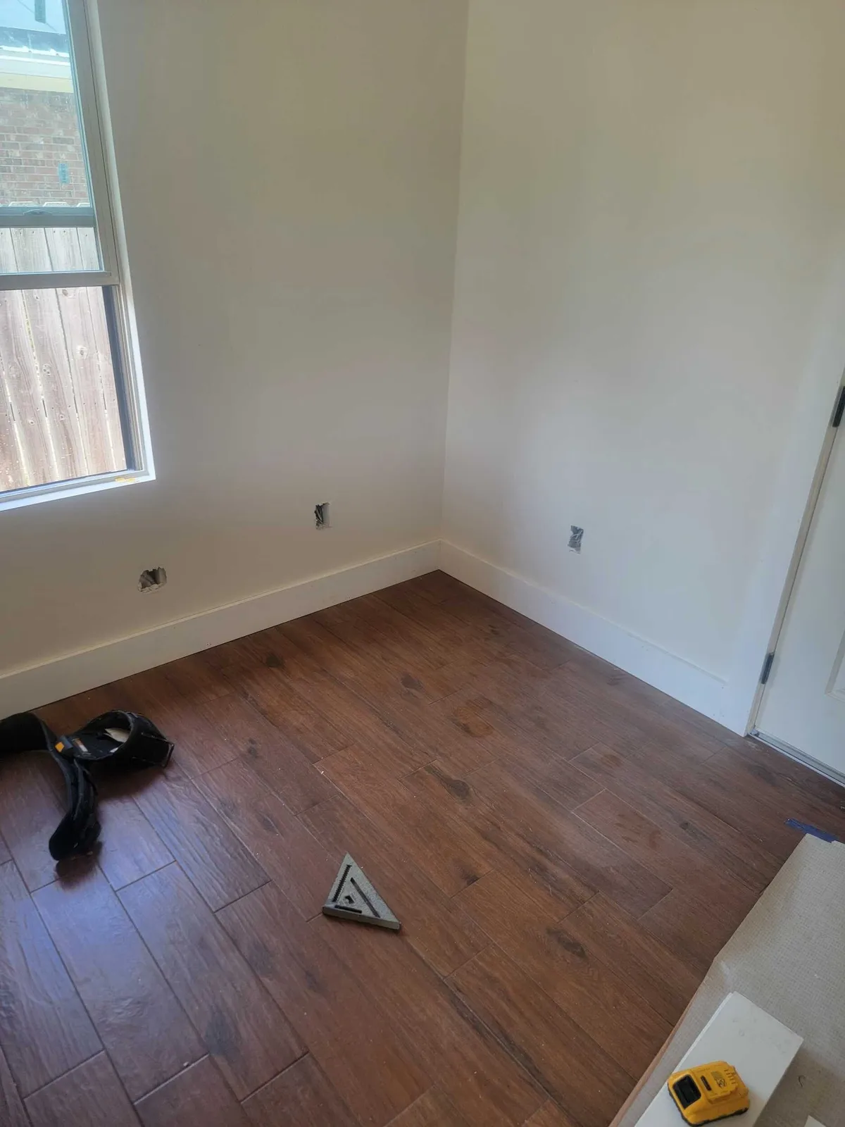 Corner of a room with wood-look plank flooring and baseboard installed, beside a window.