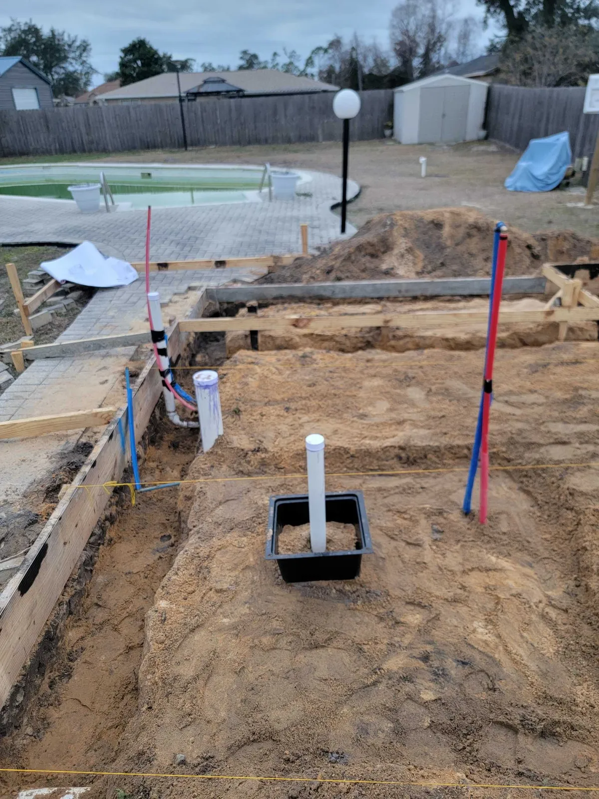 Wood foundation forms on sand with plumbing pipes rising from the ground, and a fence and pool in the background.