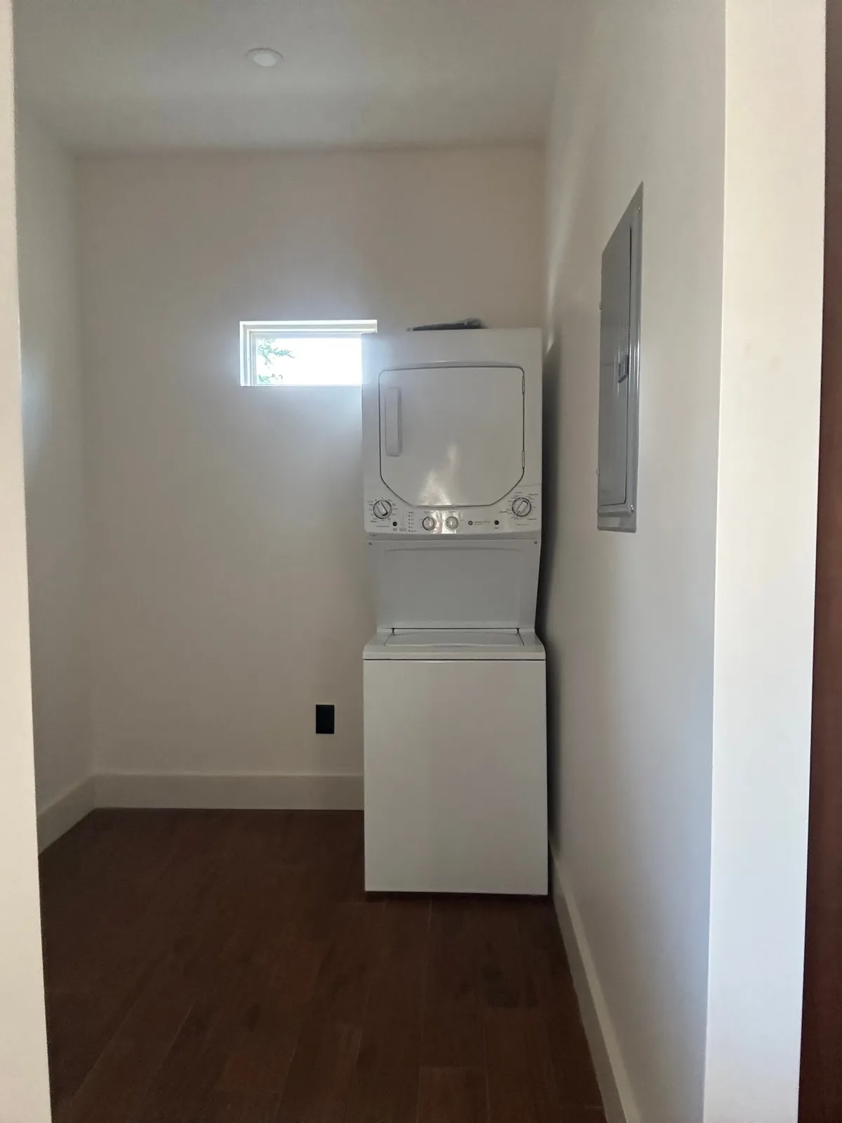 Laundry nook with a stacked washer and dryer, a small high window, and an electrical panel on the side wall.