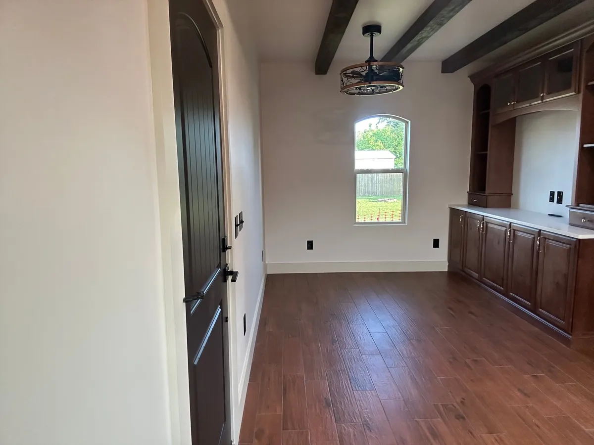 Living area with plank flooring, a wall of wood cabinetry, an arched window, a beamed ceiling with a fan, and a dark arched-top door.
