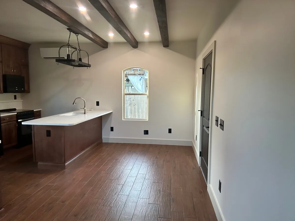Studio room with the peninsula and kitchen along one side, ceiling beams, an arched window on the far wall, and a dark door to the right.